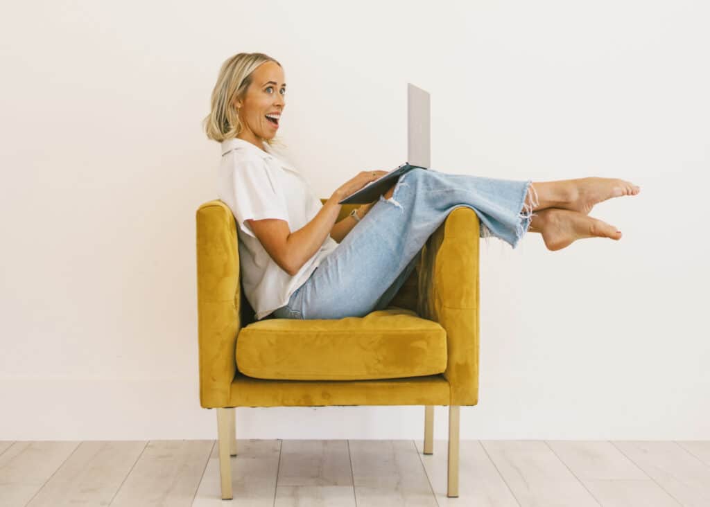 Professional travel consultant smiling while working on a laptop in a yellow armchair, representing the expert family travel coaching services of Our Family Passport.